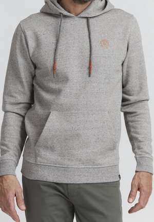 Mann trägt hellgrauen Kapuzenpullover mit Fronttasche und braunen Kordelenden, kombiniert mit dunkelgrauen Hosen.