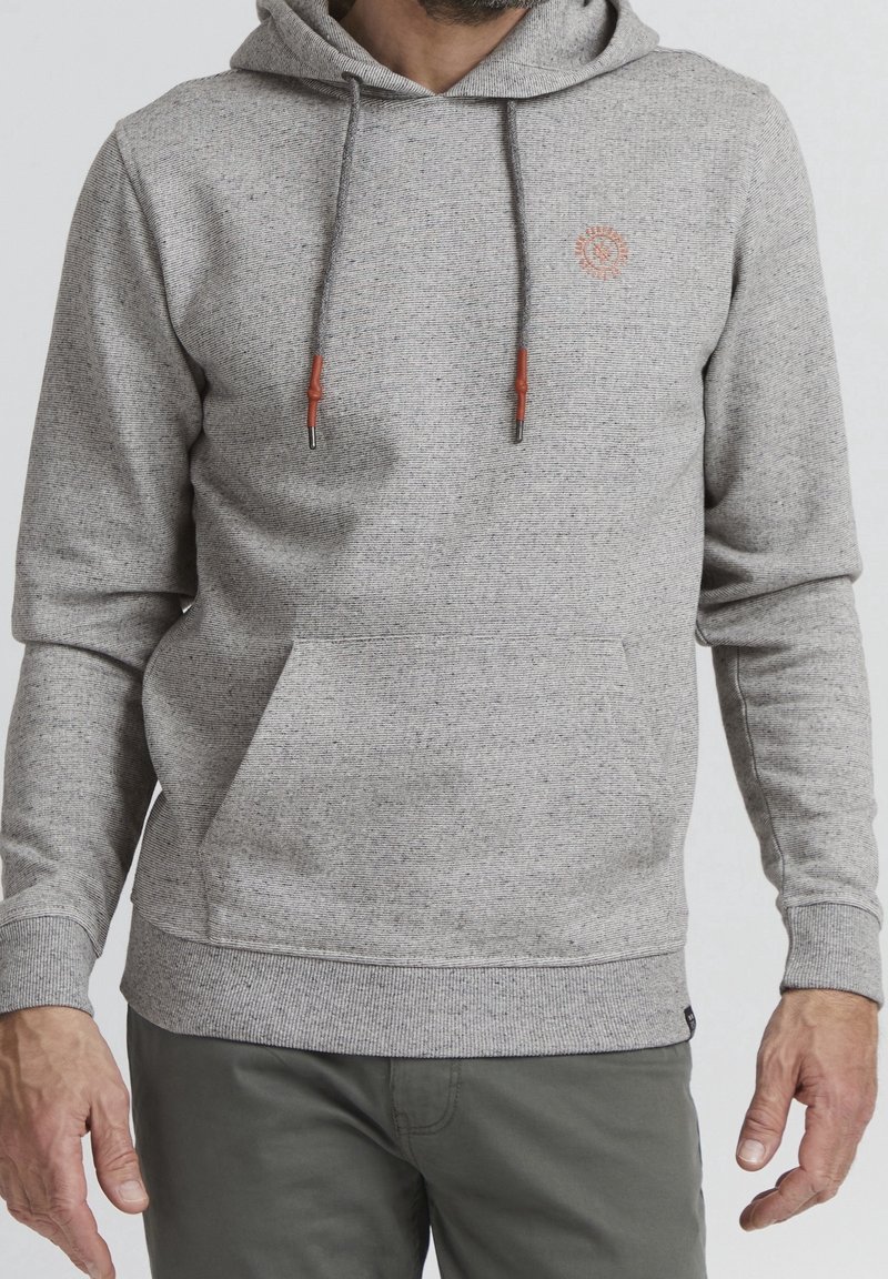 Homme portant un sweat à capuche gris clair avec poche avant et embouts de cordon marron, associé à un pantalon gris foncé.