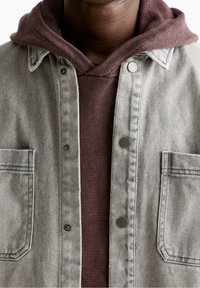 Szara kurtka dżinsowa z dwoma kieszeniami na piersi, nałożona na brązowy sweter z dzianiny. Posiada srebrne napy i teksturowany materiał.