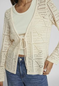 Cárdigan de punto en crema con un patrón texturizado y de tejido abierto, que cuenta con un cierre de lazo, mangas largas y un ajuste relajado sobre una blusa ligera.