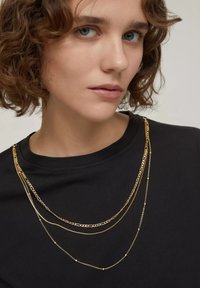 Collane sovrapposte in tono oro di diverse forme e lunghezze indossate su un capo nero, caratterizzate da catene delicate e dettagli testurizzati.