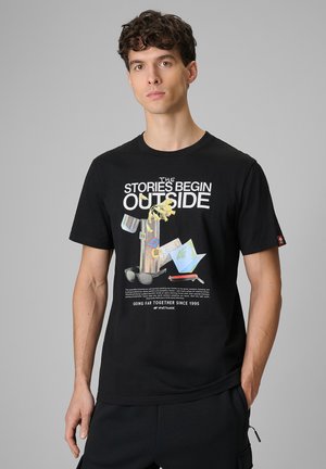 Młody mężczyzna w czarnej koszulce z grafiką o tematyce outdoor i napisem „Historie zaczynają się na zewnątrz” na jednolitym szarym tle.