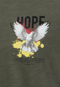 Camiseta verde oliva con una paloma blanca de alas extendidas, rodeada de salpicaduras de pintura amarilla y roja. El texto dice "ESPERO".