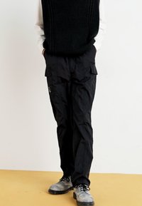 Pantalons cargo noirs avec de multiples poches, tissu léger, coupe décontractée et un détail de logo discret sur le côté, associés à des chaussures grises.