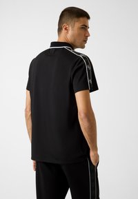 Mężczyzna stojący tyłem, ubrany w czarną koszulę polo z krótkim rękawem, z białymi wykończeniami i logo na ramionach, oraz pasujące czarne spodnie z bocznym paskiem z logo.