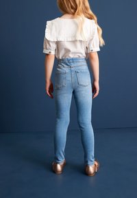 Weiße Bluse mit Rüschenschnitt, hellblaue Skinny-Jeans und glänzende bronzefarbene Schuhe. Der Hintergrund ist in einem einheitlichen dunkelblauen Farbton gehalten.