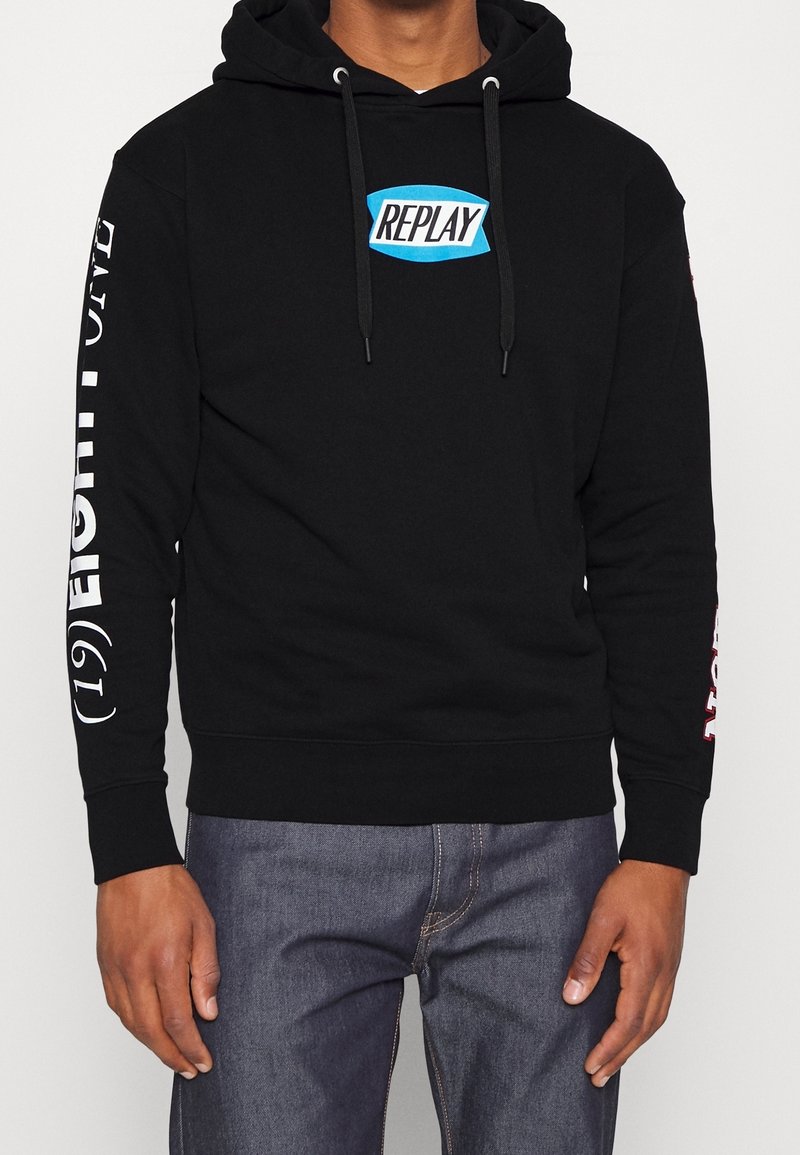 Hombre vistiendo un hoodie negro con un logo azul "REPLAY" en el pecho y texto blanco en las mangas, combinado con unos jeans azul oscuro.