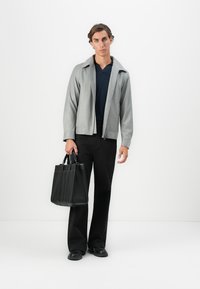 Gray zippered jacket over a navy shirt, black trousers, and black shoes. The model holds a structured black tote bag with multiple compartments.