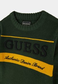 Maglione verde a maglia con colletto a coste scuro, caratterizzato da una riga orizzontale gialla con il logo "GUESS" e il testo "Authentic Denim Brand" in nero.