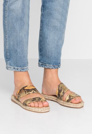 Voeten in open teen slangenprint sandalen met twee bandjes, gecombineerd met opgerolde lichtblauwe spijkerbroek op een witte vloer.
