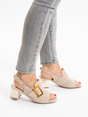 Feet wearing beige suede open-toe block heel sandals with a large rectangular buckle, paired with light gray cropped skinny jeans.