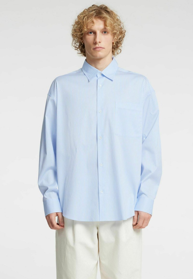 Jasnoniebieska koszula na guziki z pionowymi białymi paskami, kieszenią z przodu i długimi rękawami. Materiał wydaje się miękki i gładki. Noszona z jasnymi spodniami.