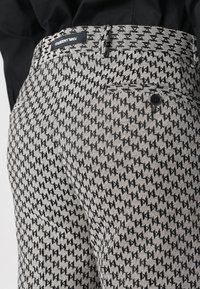 Black and white patterned pants with a geometric design, featuring a button closure, belt loops, and a branded label on the waistband.