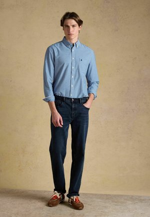 Young man standing with one hand in pocket, wearing blue checkered button-up shirt, dark jeans, and brown sneakers with orange laces.