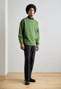 Zielony sweatshirt z ściągaczami, noszony na czarnej koszuli z kołnierzykiem. Połączony z czarnymi spodniami i masywnymi czarnymi butami, na neutralnym tle.