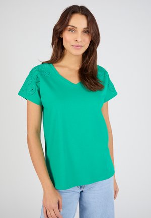 Woman wearing a green V-neck short-sleeve top with eyelet details on sleeves and light blue jeans, standing against a plain background.