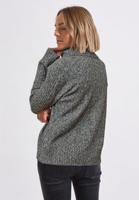 Grauer Strickpullover mit strukturiertem Muster und hohem Kragen, ausgestattet mit langen Ärmeln und einem gerippten Saum, kombiniert mit dunklen Jeans.