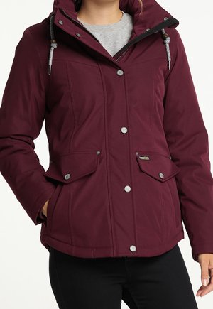 Femme portant une veste à capuche bordeaux avec boutons-pression et poches à rabat à l'avant, associée à un pantalon noir et un haut gris en dessous.