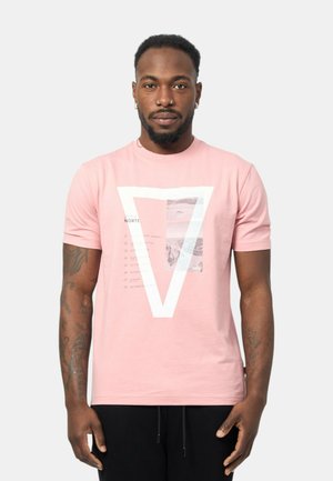 T-shirt rosa chiaro in cotone con una grafica triangolare bianca e un'inserzione fotografica. Presenta elementi testuali in basso a sinistra. Maniche corte.