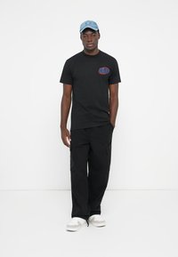 Sort kortærmet T-shirt med et rødt og blåt "HUF"-logo, kombineret med sorte cargo-bukser og en lyseblå kasket. Hvide sneakers fuldender outfittet.