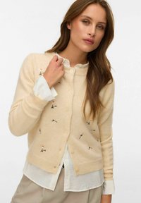 Žena s dlouhými hnědými vlasy, která má na sobě béžový rozepínací svetr s malou květinovou výšivkou přes bílou blůzu a béžové kalhoty.