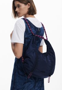 Hobo-Tasche aus dunkelblauem Nylon mit gerafften Details, lila-goldener Seiltrageriemen, getragen über einem Jeanskleid, das die weiche Textur der Tasche zur Geltung bringt.