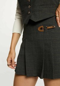 Donna che indossa una gonna a scacchi scuri con pieghe e dettaglio in pelle con fibbia, abbinata a un gilet coordinato e a una maglietta a maniche lunghe color crema.