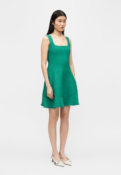 Femme debout portant une robe verte sans manches côtelée avec un col carré et des escarpins slingback crème à bouts pointus, sur un fond blanc.