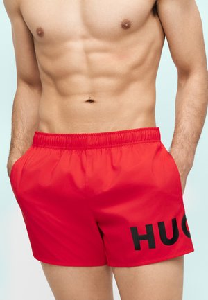 Shirtless man with muscular torso wearing red swim shorts with "HUGO" logo, hands resting in pockets against plain background.