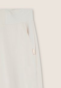 Pantalon de jogging beige clair avec une ceinture tissée texturée, poche latérale avec des accessoires en or rose et surface en tissu lisse.