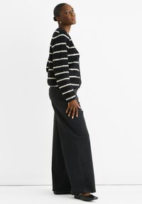 Schwarz-weiße gestreifte Strickjacke mit Knopfverschluss, kombiniert mit weit geschnittenen schwarzen Jeans und schwarzen Ballerinas, von der Seite gezeigt.