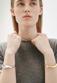 Brazaletes de oro y plata, de diseños circulares y retorcidos, llevados en las manos. Parte superior corta de manga gris. El cuello del modelo adornado con una cadena de oro.