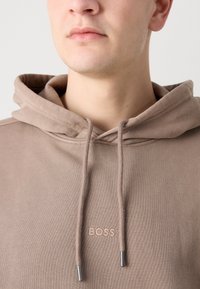 Bruine hoodie met een trekkoord, voorzien van zilveren hardware en een subtiele "BOSS" logo in lichte stiksels. Zachte, gestructureerde stof.