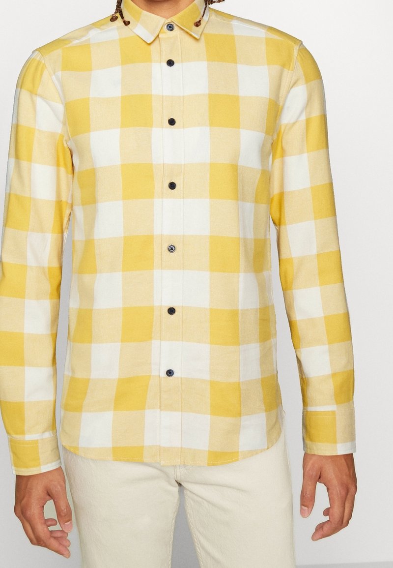 Camisa de cuadros amarillos y blancos con mangas largas, cuello abotonado y botones negros. Textura suave, corte regular y diseño de dobladillo curvado.