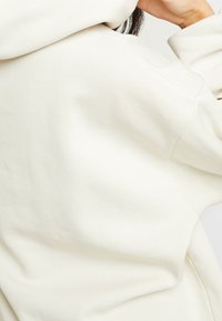 Kremowa bluza z kapturem wykonana z miękkiego materiału; posiada duży kaptur i luźny fason; widoczne przeszycia oraz detale faktury na powierzchni.