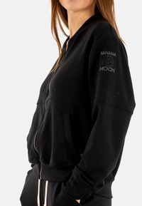 Chaqueta bomber negra con cremallera, bolsillos laterales, puños acanalados y un logo en la manga. Fabricada con un tejido suave y liso.