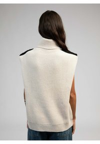 Pull sans manches en tricot couleur blanc cassé avec un col montant, présentant des accents d'épaule noirs et une texture côtelée.