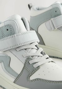 Sneakers albe și gri deschis, cu un aspect din piele netedă, cu guler căptușit, curea tip Velcro și talpă din cauciuc texturată cu perforații.
