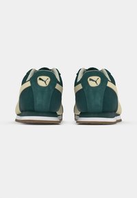 Πράσινα σουέτ αθλητικά παπούτσια με κρέμα λεπτομέρειες, που διαθέτουν λευκή σόλα και καφέ σόλα. Το λογότυπο Puma είναι ορατό στην φτέρνα και στη γλώσσα.