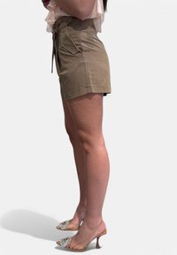 Shorts in khaki a vita alta con cintura da annodare, tasche laterali e taglio dritto, abbinati a sandali con tacco trasparente e dettaglio di gioiello.