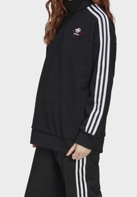 Svart fleece-pullover med hög krage, vitt randiga ärmar och röd logodetalj. Har en avslappnad passform och ribbade ärmslut.