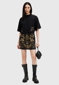 Svart cropped t-skjorte med gullbroderi, kombinert med et svart mini skjørt dekket av paljetter med intrikate gullmønstre og svarte høye støvler.