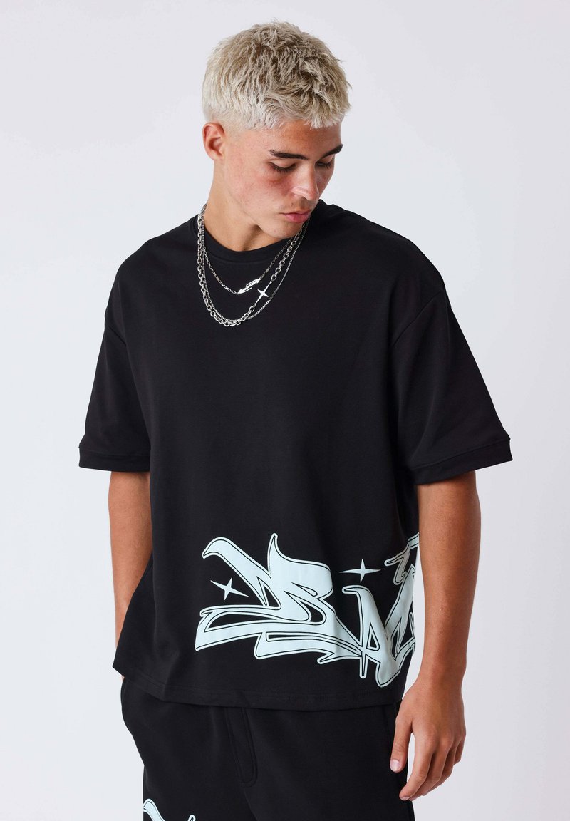 Giovane uomo con capelli biondi corti che indossa una T-shirt nera oversize con grafica bianca in stile graffiti vicino all'orlo, che guarda verso il basso.