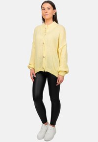 Cardigan en maille surdimensionné jaune avec une finition texturée, doté de grands boutons, porté sur des leggings noirs et des baskets blanches.