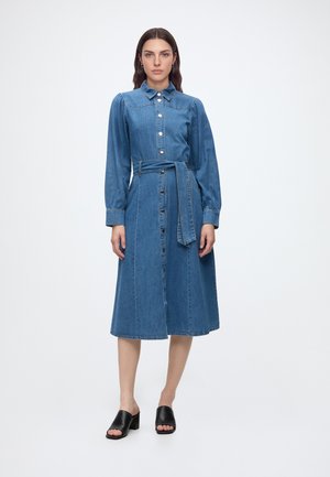 Femme debout portant une robe midi en denim bleu avec des manches longues, des boutons sur le devant, une ceinture en tissu et des sandales noires à talons ouvertes.