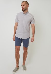 Camisa de botones de manga corta en gris claro con cuello, combinada con pantalones cortos azul marino y zapatos gris claro tipo slip-on, sobre un fondo liso.