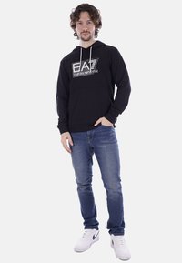 Felpa nera con cappuccio realizzata in tessuto morbido, dotata di tasca a marsupio anteriore e cappuccio con cordino, caratterizzata da un logo bianco a contrasto. Indossata con jeans blu.
