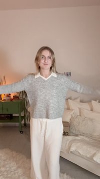 Gray knitted sweater with a V-neck over a white collared shirt, paired with white loose-fitting pants. Cozy texture in a home setting.