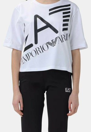 Persona che indossa una t-shirt corta bianca EA7 Emporio Armani e pantaloni neri con il logo EA7 Emporio Armani sulla coscia sinistra.