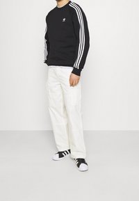 Svart sweatshirt med vit tre-stripes design på ärmarna och logotyp, tillsammans med vita cargobyxor och svart-vita sneakers.